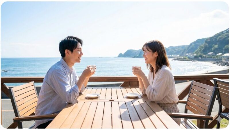 晴れた日の海沿いのカフェテラスで、笑顔で見つめ合いながらコーヒーを楽しむ日本人カップル。ドライブデートのおすすめスポットの様子