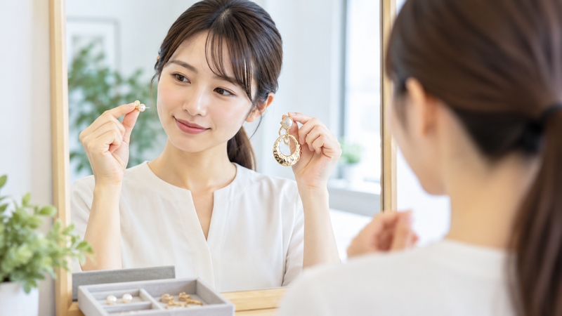 小ぶりなピアスと大ぶりなアクセサリーを鏡の前で見比べる女性