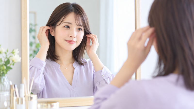 明るい色のVネック風ブラウスを着て髪を整えながら鏡を見る女性